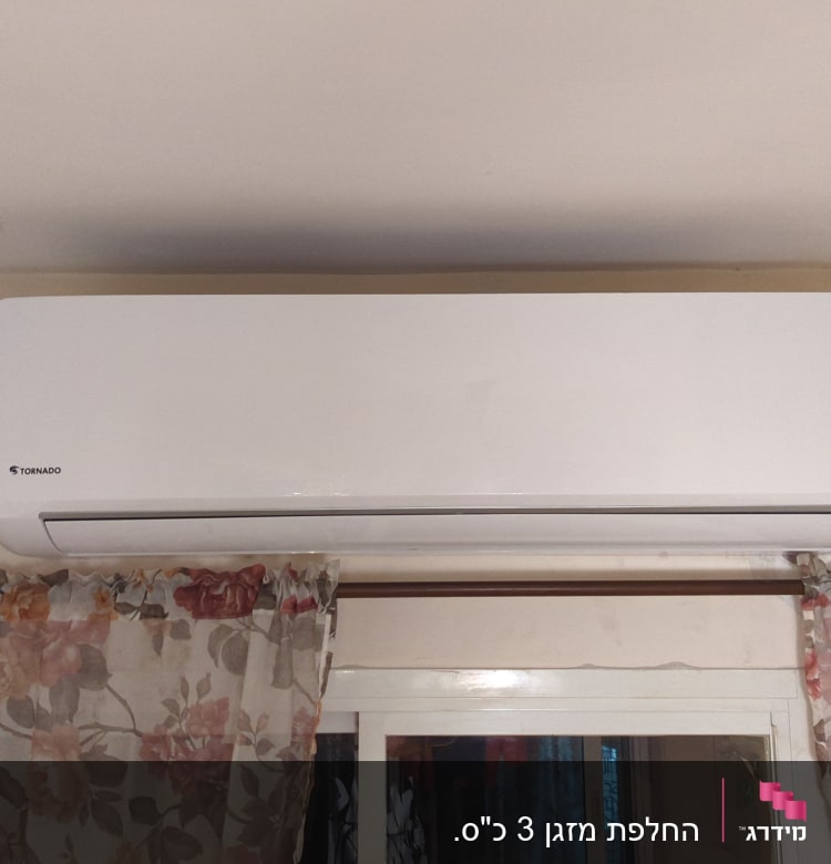 מזגן קיר לבן מותקן מעל חלון עם וילון פרחוני